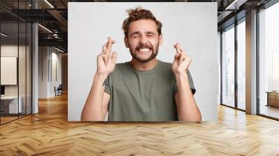 Cheerful bearded man keeps fingers crossed, smiles broadly and closes eyes, makes desire wish. Positive male student believes in victory or success on exam. Feelings, attitudes and reactions Wall mural