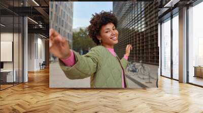 Cheerful carefree woman strolls in city and dances keeps arms sideways enjoys good day walks in downtown among modern city buildings wears jacket being in good mood. People leisure emotions. Wall mural