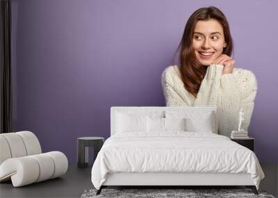 Cheerful Caucasian girl keeps hands together near face, looks positively aside, has no make up, healthy skin, wears white sweater, stands over purple background with blank space for your promotion Wall mural