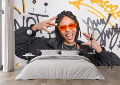 Cheerful cool ethnic woman with dreadlocks makes yo gesture has fun dressed in black jacket and stylish orange sunglasses smiles broadly poses against graffiti background. Youth subculture concept Wall mural