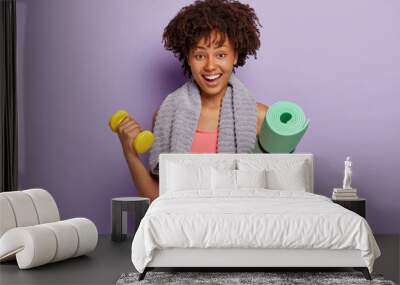 Cheerful dark skinned young Afro woman holds kareamt and dumbbell, trains muscles in gym, has happy facial expression, towel around neck, wears pink top, models indoor against purple background Wall mural