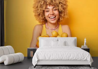 Cheerful European woman with blonde curly hair embraces heap of fresh juicy tropical fruits eats healthy food dressed in casual t shirt isolated over vivid yellow background. Monochrome shot Wall mural