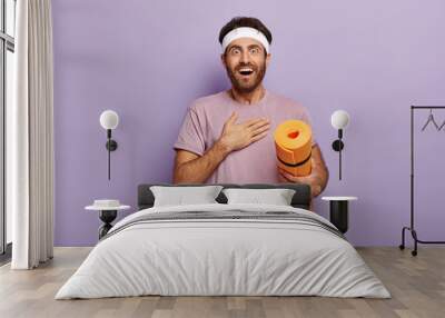 Cheerful excited sportsman looks with surprisement, touches chest, practices yoga every day, being in good physical shape, dressed in sportwear, holds orange fitness mat, isolated on purple wall Wall mural