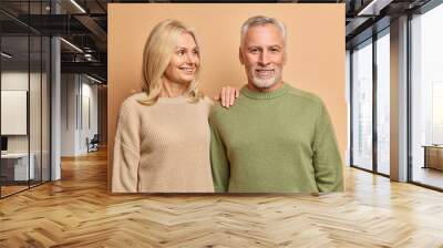 Cheerful mature couple stand next to each other have happy expressions demonstrate love exists in old age going to celebrate anniversary of wedding pose indoor against beige studio background Wall mural