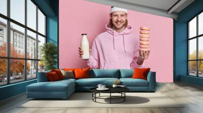 Cheerful sportsman holds bottle of milk and pile of doughnuts has temptation to eat junk food smiles happily wears headband and hoodie isolated over pink background. Sport and nutrition concept Wall mural