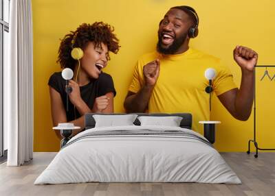 Cheerful young African American bloggers enjoy favourite playlist in headphones, listens audio in music app, feels happiness, dance actively against yellow background, move emotionally, have fun Wall mural