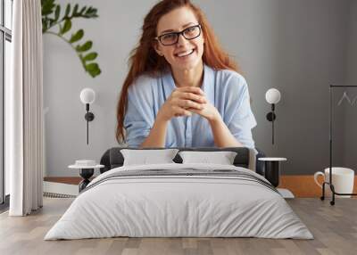 Cheerful young businesswoman in casual shirt and glasses looking and smiling at the camera. Happy successful female entrepreneur enjoying productive day while sitting in front of the computer Wall mural