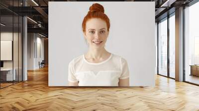 Cheerful young Caucasian woman with freckles and ginger hair in bun looking and smiling happily at camera, wearing white blouse. Redhead girl with pretty joyful smile posing at blank studio wall Wall mural