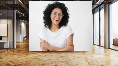 Cheerful young good looking woman with clean dark skin and black shaggy hair posing indoors with crossed arms, smiling broadly with her white straight teeth, laughing at good joke, wearing casual top Wall mural