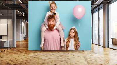 Children and celebration concept. Bearded unshaven man gives piggy back to small daughter, long haired girl stands near and holds pink balloon, have family party birthday day, have fun indoors Wall mural