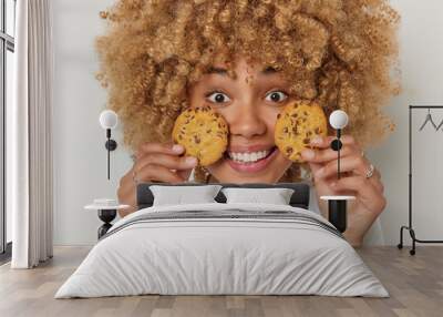 Close up portrait of cheerful woman with curly bushy hair holds cookies near face breaks diet has glad facial expression dressed in casual t shirt isolated over white background. Unhealthy food Wall mural