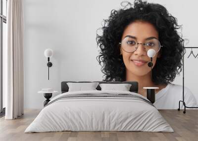 Close up portrait of curly female adult has charming smile, curly dark hair, wears big glasses, satisfied as finished domestic work earlier, being successful designer or architect, has talent Wall mural