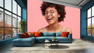 Close up portrait of happy African American female laughs at something funny, has positive expression, wears glasses, has curly hair, dressed casually, closes eyes with happiness, isolated on pink Wall mural