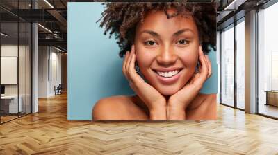 Close up portrait of relaxed black woman has gentle skin after taking shower, satisfied with new lotion, has no makeup, smiles tenderly, shows perfect teeth, stands shirtless against blue background Wall mural