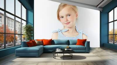 Close up shot of beautiful blonde Caucasian little girl wearing striped dress, looking at the camera with adorable smile, posing against white copy space concrete wall. Happy childhood concept Wall mural