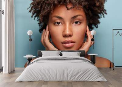 Close up shot of black young woman with Afro hairstyle, touches cheeks with both hands, has bare shoulders, tender look isolated over blue background, looks directly at camera. Horizontal shot Wall mural