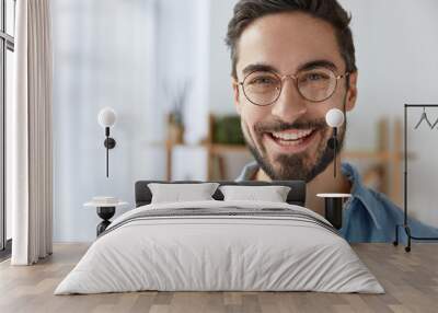 close up shot of cheerful satisfied attractive male with stubble, has broad smile, wears round spect Wall mural