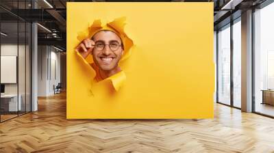 Close up shot of glad Caucasian man keeps hand on rim of spectacles, wears hat in one tone with background, looks happily through torn paper hole, copy space on right side for your promotional content Wall mural