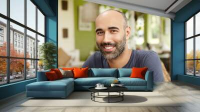 Close up shot of handsome Caucasian man with beard dressed in t-shirt looking and smiling at camera with happy cheerful expression, sitting at sidewalk restaurant on sunny day, waiting for friends Wall mural