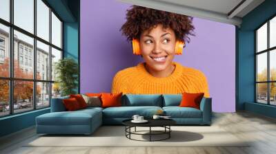 Close up shot of happy dark skinned woman listens music via headphones bites lips and enjoys pleasant melody dressed in knitted orange sweater poses against purple background. My favorite tune Wall mural