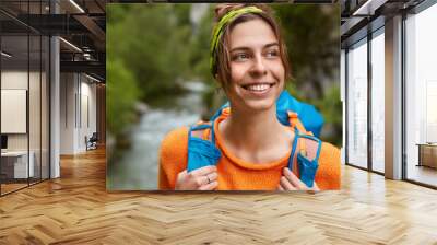 Close up shot of mirthful positive Caucasian female looks away, spends summer vacation actively in nature, carries backpack, dressed in orange jumper, poses against landscape, feels free and delighted Wall mural