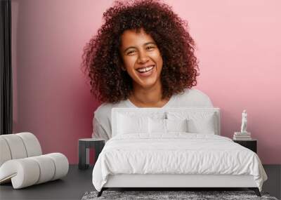 Close up shot of pretty teenage girl with dark skin, curly Afro hair, grins at camera, has white teeth, laughs sincerely at good joke, has fun with close friend, wears everyday white t shirt Wall mural