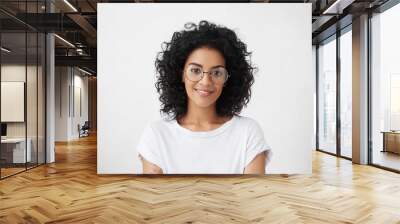 Close up studio shot of beautiful young mixed race woman model with curly dark hair looking at camera with charming cute smile while posing against white blank copy space wall for your content Wall mural