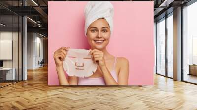 Complexion treatment concept. Young pretty woman holds gel peel mask for face, has cheerful expression, wears towel on head after taking bath, dressed in casual wear, poses over pink background Wall mural