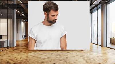 Concentrated handsome young male focused down, has dark stubble and hair, wears casual white t shirt, isolated over studio background with copy space for your advetisement or promotional text Wall mural