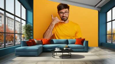 Confident charismatic man makes call me gesture, communicates with gestures, asks girlfriend telephone number, looks with flirty expression, isolated on yellow wall, wears casual outfit. Body language Wall mural