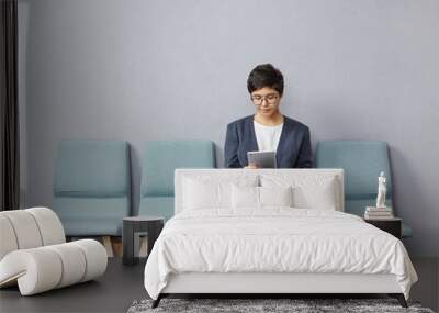 Confident successful young businesswoman with short hairstyle wearing stylish clothes and glasses using digital tablet while waiting for doctor's appointment, sitting on chair in hall at clinic Wall mural