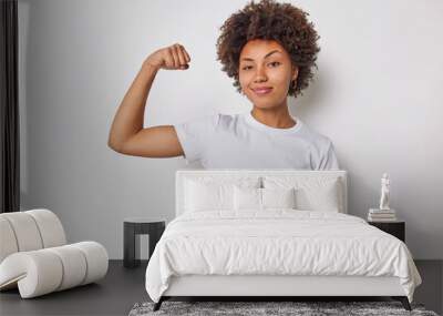 Confident woman feels strength and power raises arm flexes biceps and looks proud of her own achievements has strong muscles wears casual t shirt isolated over white background brags with fit body Wall mural