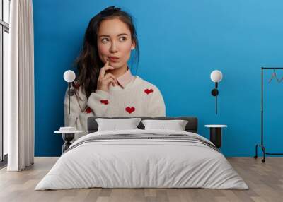 Contemplative pretty millennial young Asian woman looks away, builds plans in mind, has thoughtful face expression, long dark pony tail, wears sweater with hearts, isolated over blue background Wall mural