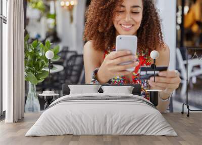 Cropped shot of beautiful African American woman with happy look, holds modern cell phone and credit card, makes shopping online, drinks smoothie in outdoor cafeteria. People and payment concept Wall mural