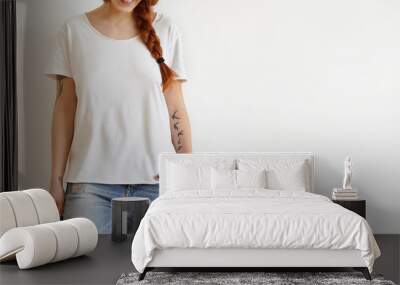 Cropped shot of beautiful stylish young redhead woman with braid and tattoos smiling joyfully dressed in white oversize t-shirt, keeping hand in pocket of her trendy blue ragged jeans. Horizontal Wall mural