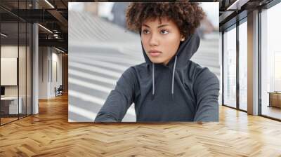 Cropped view of thoughtful dark skinned young sportswoman in stylish hoodie, focused into distance, sits on road against blurred background, has healthy skin. Ethnicity and lifestyle concept Wall mural