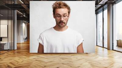 Curious intelligent male wears round spectacles, casual t shirt, notices something down, concentrated on floor, has puzzled look, isolated over white studio background. People and facial expressions Wall mural