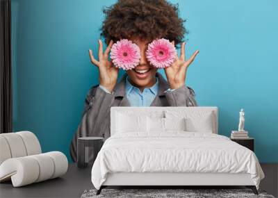 Curly haired positive woman keeps pink gerbera daisy in front of eyes, hides face and smiles broadly. Positive Afro American lady poses with flowers against blue wall. Female gardener indoor Wall mural