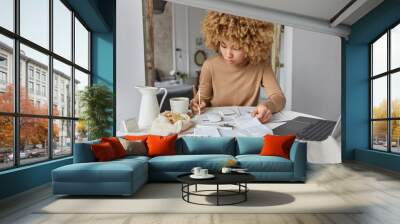 Curly haired woman sits at desk at home manages household budget studies financial bills and expenditures wears casual brown jumper plans expenses poses against domestic interior. Paper work concept Wall mural