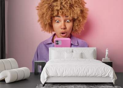 Curly haired young woman stares in smartphone with widely opened mouth and bugged eyes reads shocking news receives unexpected message dressed in purple jacket isolated over pink background. Wall mural