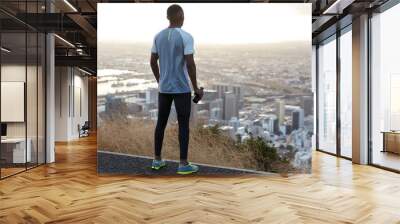 Dark skinned sportsman in sportswear, stands back, drinks water from bottle, wears sneakers, stands high and enjoys scenic city view with sky scrapers from above, has outdoor workout in countryside Wall mural