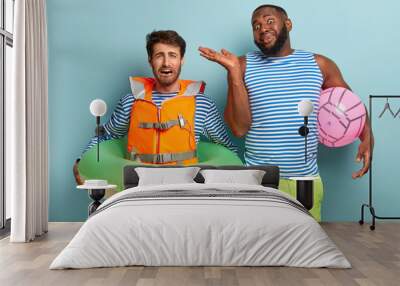 Dejected man tries to swim, wears lifevest, poses with inflated swimring. Funny unaware black guy in sailor vest and green shorts, holds beach ball. Mixed race male friends spend summer time on sea Wall mural