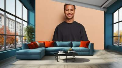 Delighted teenager with dark skin stands cheerful man smiles pleasantly dressed in casual black t shirt poses against brown background listens something with pleased expression. Studio shot. Wall mural