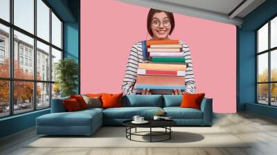 Delighted young woman carries pile of textbooks, smiles broadly, learns useful information from encyclopedia, has dark hair, dressed in casual outfit, poses aginst pink background. Education concept Wall mural