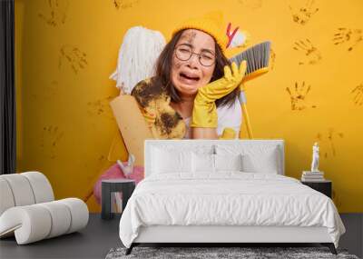 Desperate frustrated housewife cries unhappily holds dirty sponge wears hat rubber gloves poses with cleaning equipment does laundry at home expresses negative emotions isolated on yellow wall Wall mural