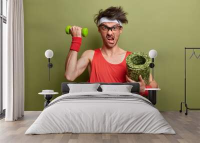 Determined sporty man poses with dumbbell and rolled up karemat, ready for workout, looks with crazy expression, wears headband, shirt and wristbands, isolated on green background works on arm muscles Wall mural