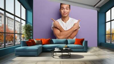 Different direction and hesitation. Unsure attractive man crosses hands over chest, points to both sides, right and left, makes choice between two items, wears casual t shirt, isolated on purple wall Wall mural