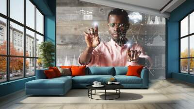 Double exposure. Black young businessman wearing glasses and casual shirt, touching futuristic screen interface with his fingers, making calculations, analyzing diagrams and schemes. Selective focus Wall mural