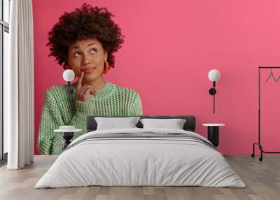 Dreamy dark skinned young woman thinks about career opportunities, stands thoughtful, concentrated upwards, dressed in knitted sweater, isolated on pink background, copy space for your promotion Wall mural