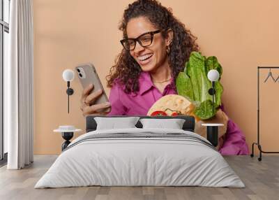 Eat healthy food. Pleased Brazilian female model with curly hair holds paper grocery bag makes shopping online via smartphone returns from market isolated over brown background. Delivery concept Wall mural